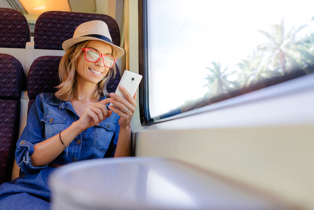 Fiatal, farmerruhát viselő nő vonaton, mobiltelefonnal, szalmakalapban és napszemüvegben