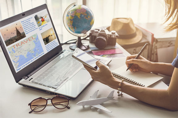A képen egy íróasztal látható, rajta egy földgömb, egy laptop, egy szemüveg, egy repülőgépmakett, és egy mobiltelefont tartó, jegyzetfüzetbe író nő két keze látható.